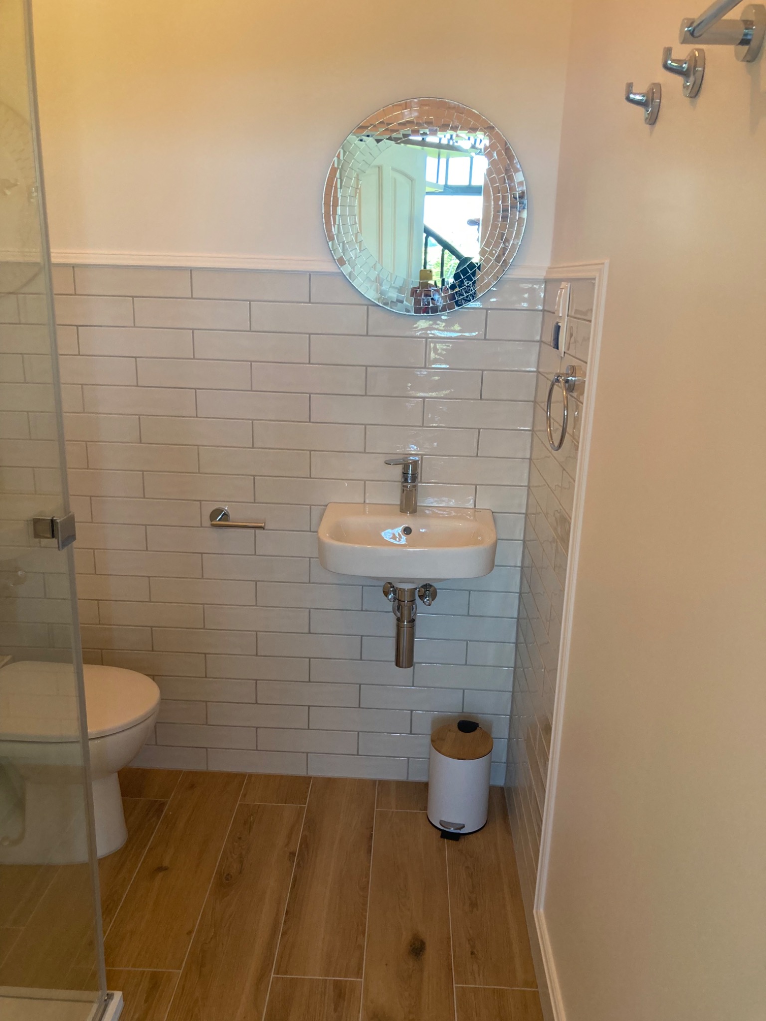 Mała łazienka z beżowymi płytkami imitującymi cegłę, drewnopodobną podłogą, okrągłym lustrem z mozaiką, białą umywalką i toaletą za szklaną kabiną prysznicową.