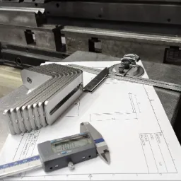 Stos precyzyjnie wykonanych, kątowych elementów z metalu, ułożonych na rysunku technicznym z wymiarami, obok kątomierza i suwmiarki elektronicznej, na tle metalowej konstrukcji.