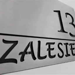 Metalowa tabliczka z wyciętym numerem domu '13' i nazwą 'ZALESIE', zamocowana na ścianie.