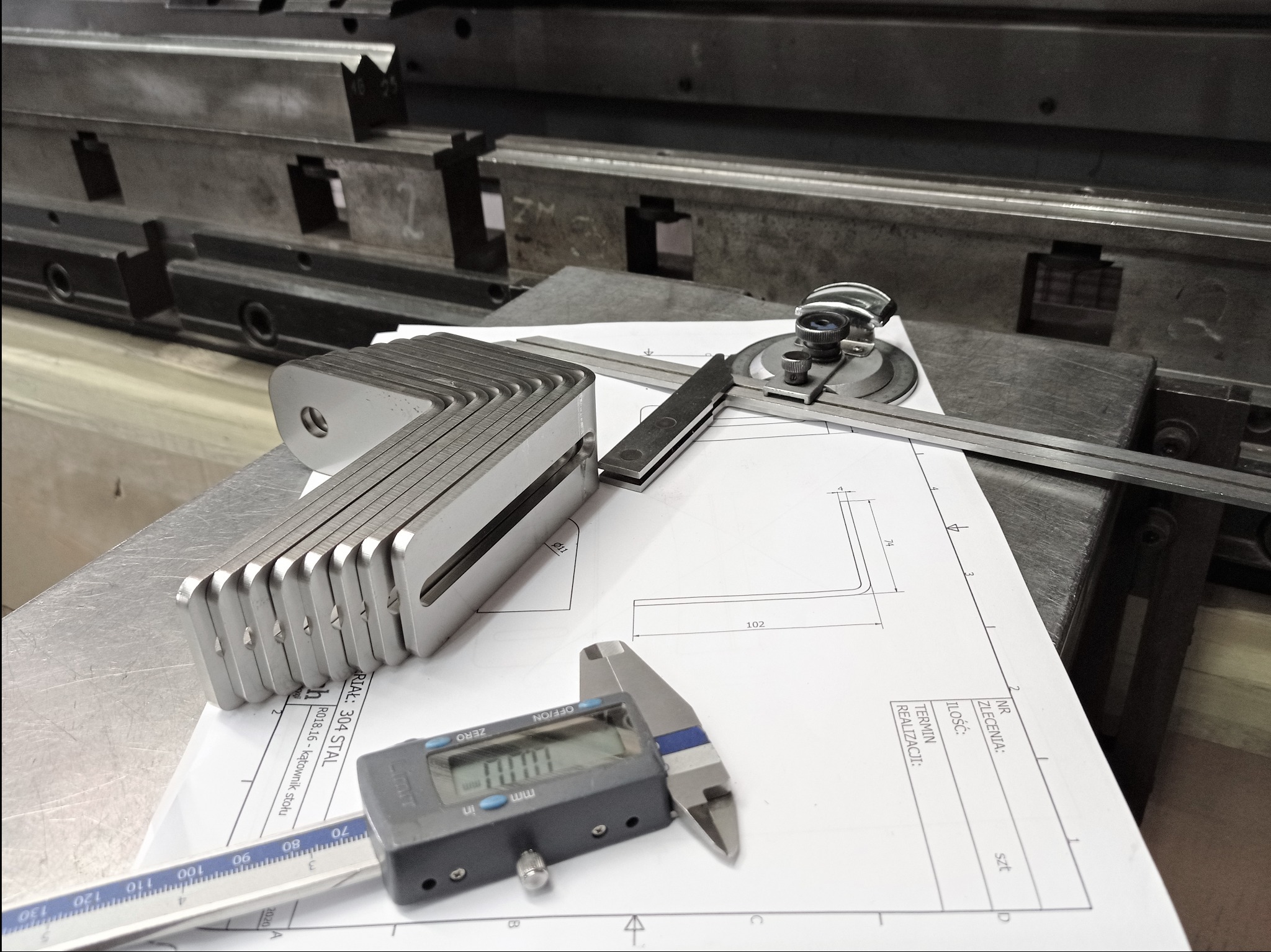 Stos precyzyjnie wykonanych, kątowych elementów z metalu, ułożonych na rysunku technicznym z wymiarami, obok kątomierza i suwmiarki elektronicznej, na tle metalowej konstrukcji.