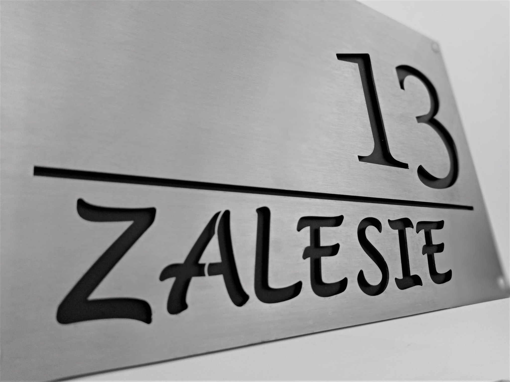 Metalowa tabliczka z wyciętym numerem domu '13' i nazwą 'ZALESIE', zamocowana na ścianie.