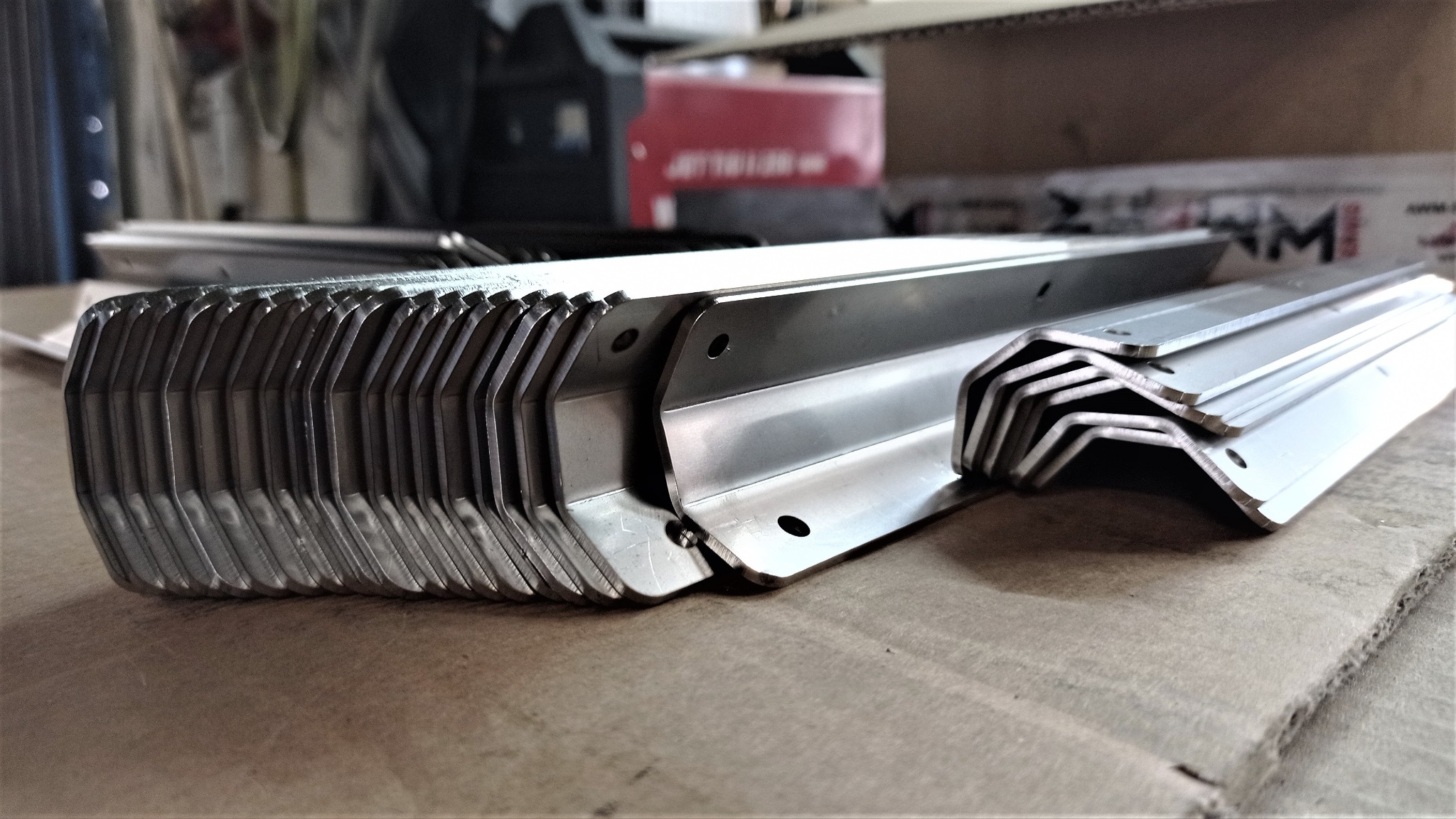 Stos wyciętych i uformowanych metalowych kątowników z otworami, ułożonych na tekturowej powierzchni roboczej.