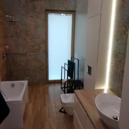 Wykończona łazienka z brązowymi płytkami imitującymi kamień, drewnopodobną podłogą, białą wanną, umywalką nablatową i oświetleniem LED.