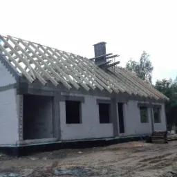 Surowy stan domu jednorodzinnego w trakcie budowy: ściany z betonu komórkowego, konstrukcja dachu z drewna, komin z cegły, widoczne otwory okienne i drzwiowe, teren wokół w stanie roboczym.