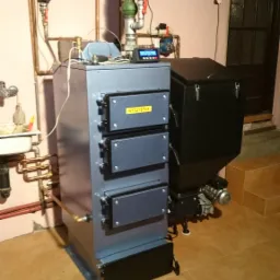 Szary piec na paliwo stałe z podajnikiem, widoczne rury miedziane, sterownik elektroniczny, kluczyk do czyszczenia na podłodze z płytek.