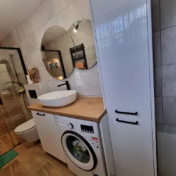 Nowoczesna łazienka z umywalką nablatową na drewnianym blacie, pod którą znajduje się pralka, obok wysoka, biała szafka, na ścianie dwa okrągłe lustra i szare płytki imitujące kamień.