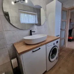 Nowoczesna łazienka z drewnianym blatem, na którym umieszczona jest owalna umywalka i czarna bateria. Obok zabudowana pralka i wysoka biała szafka. W lustrze odbija się lampa sufitowa i okno...