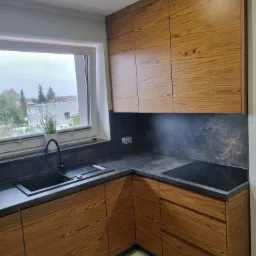 Nowoczesna kuchnia z drewnianymi frontami szafek, czarnym blatem i zlewozmywakiem, oraz płytą indukcyjną, z widokiem na zabudowę miejską zza okna.