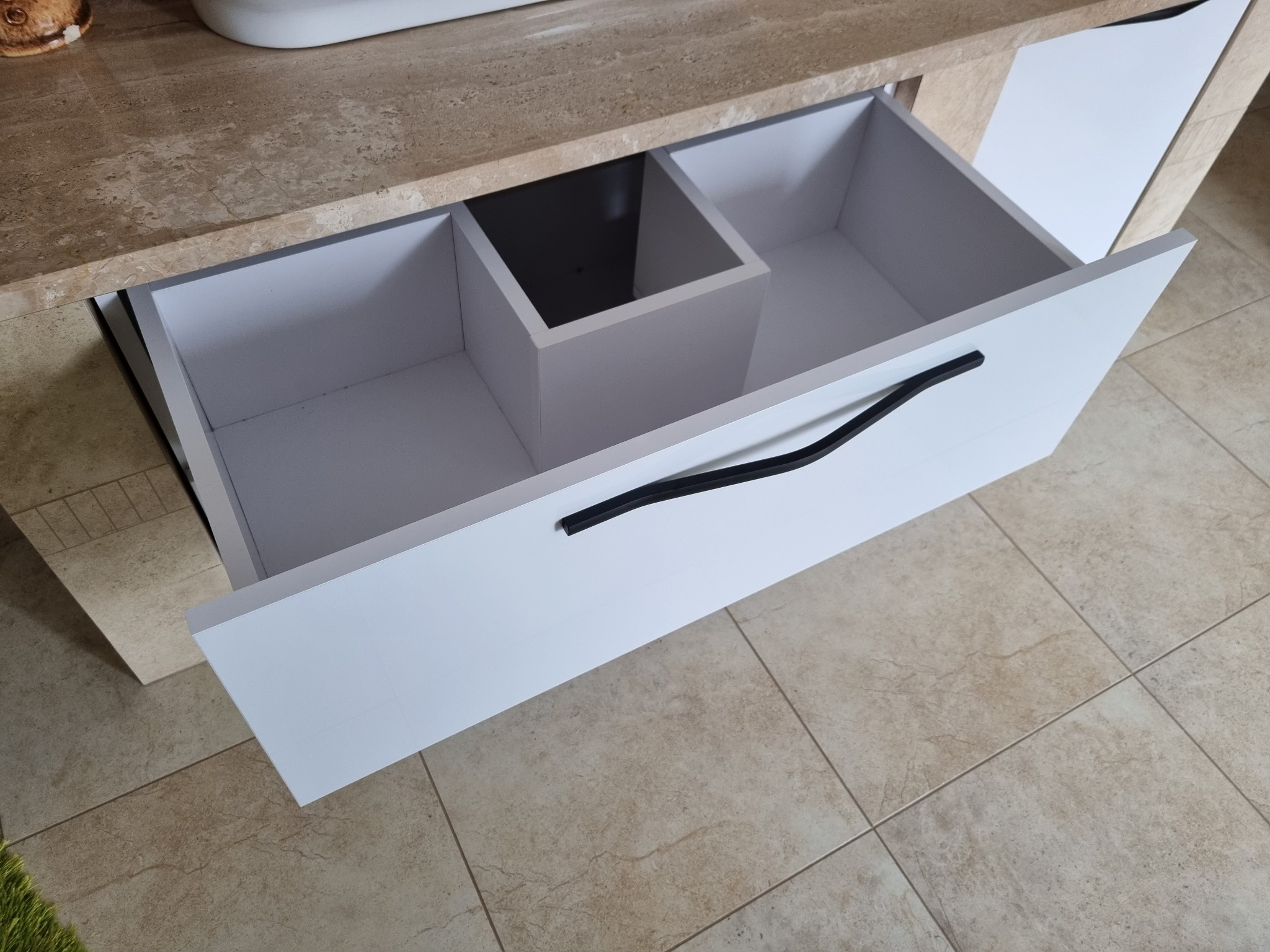 Otwarta biała szuflada z czarnym uchwytem w szafce z blatem imitującym kamień, widoczne wewnętrzne przegródki szuflady.