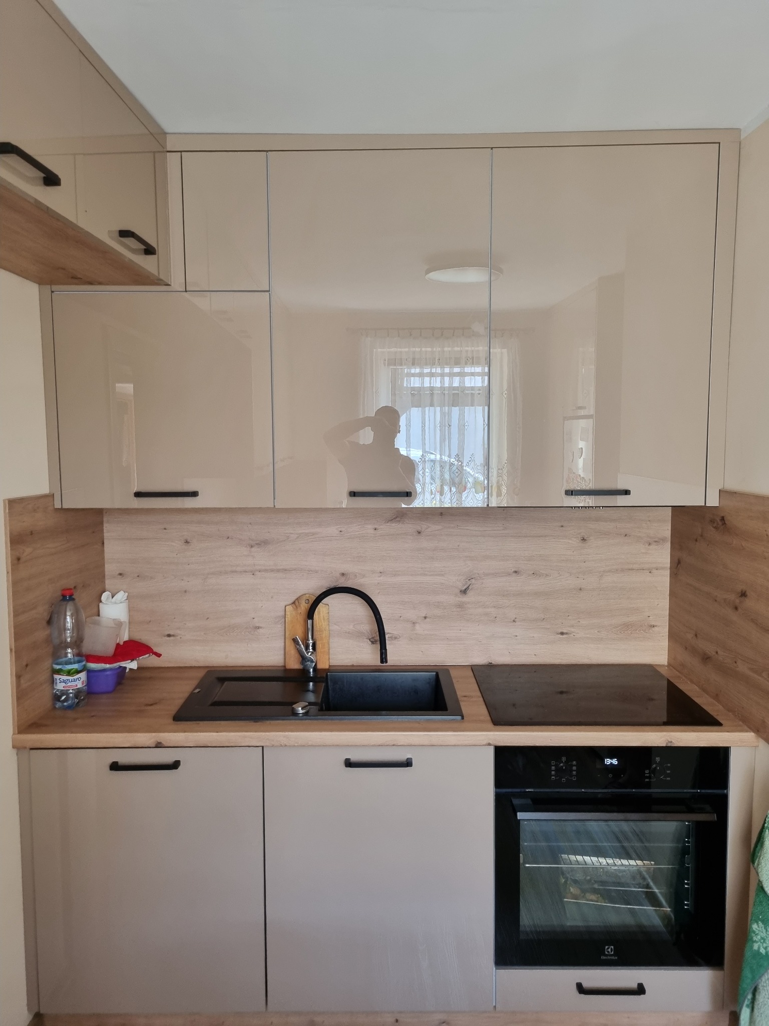 Nowoczesna kuchnia z beżowymi, lakierowanymi frontami szafek, drewnopodobnym panelem ściennym, czarnym zlewem i płytą indukcyjną, widoczny odbijający się w szafkach zarys sylwetki osoby.