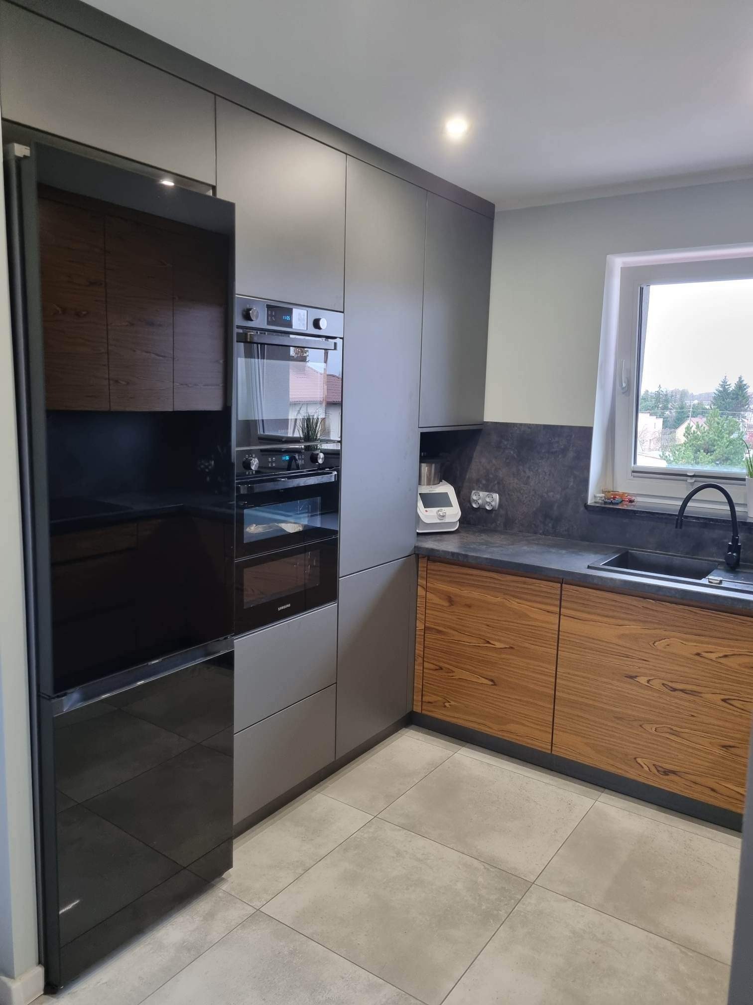 Nowoczesna kuchnia z szarymi i drewnianymi frontami, czarnym sprzętem AGD i blatem, jasne płytki na podłodze, widok na zewnątrz przez okno.