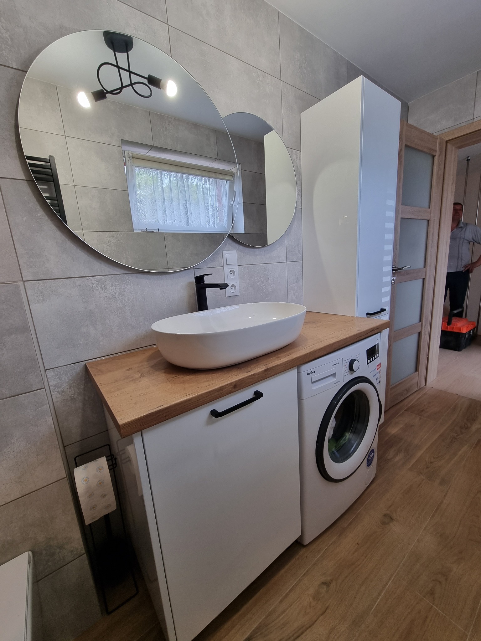Nowoczesna łazienka z drewnianym blatem, na którym umieszczona jest owalna umywalka i czarna bateria. Obok zabudowana pralka i wysoka biała szafka. W lustrze odbija się lampa sufitowa i okno...