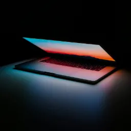 Otwarty laptop na ciemnym tle, ekran wyświetla krajobraz o zachodzie słońca, klawiatura podświetlona na czerwono.