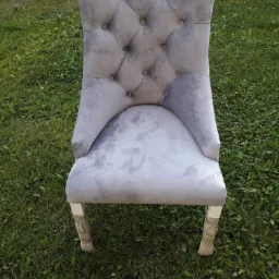 Szary, tapicerowany krzesło z pikowanym oparciem w stylu glamour, stojące na trawie. Na materiale widoczne powtarzające się logo producenta.