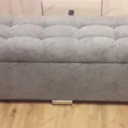 Szara, tapicerowana pufa z pikowanym siedziskiem na niskich, metalowych nóżkach, na tle drewnianej podłogi i mebli.