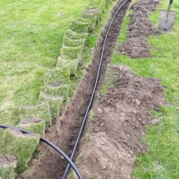 Wykopany wąski rów w trawniku z ułożoną czarną rurą, obok wykopane kostki darni i pryzma ziemi, widoczne narzędzia ogrodnicze w tle.