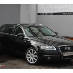 Czarny samochód Audi A6 Avant zaparkowany w salonie samochodowym, widoczne logo dealera na grillu i numer telefonu nad nim.