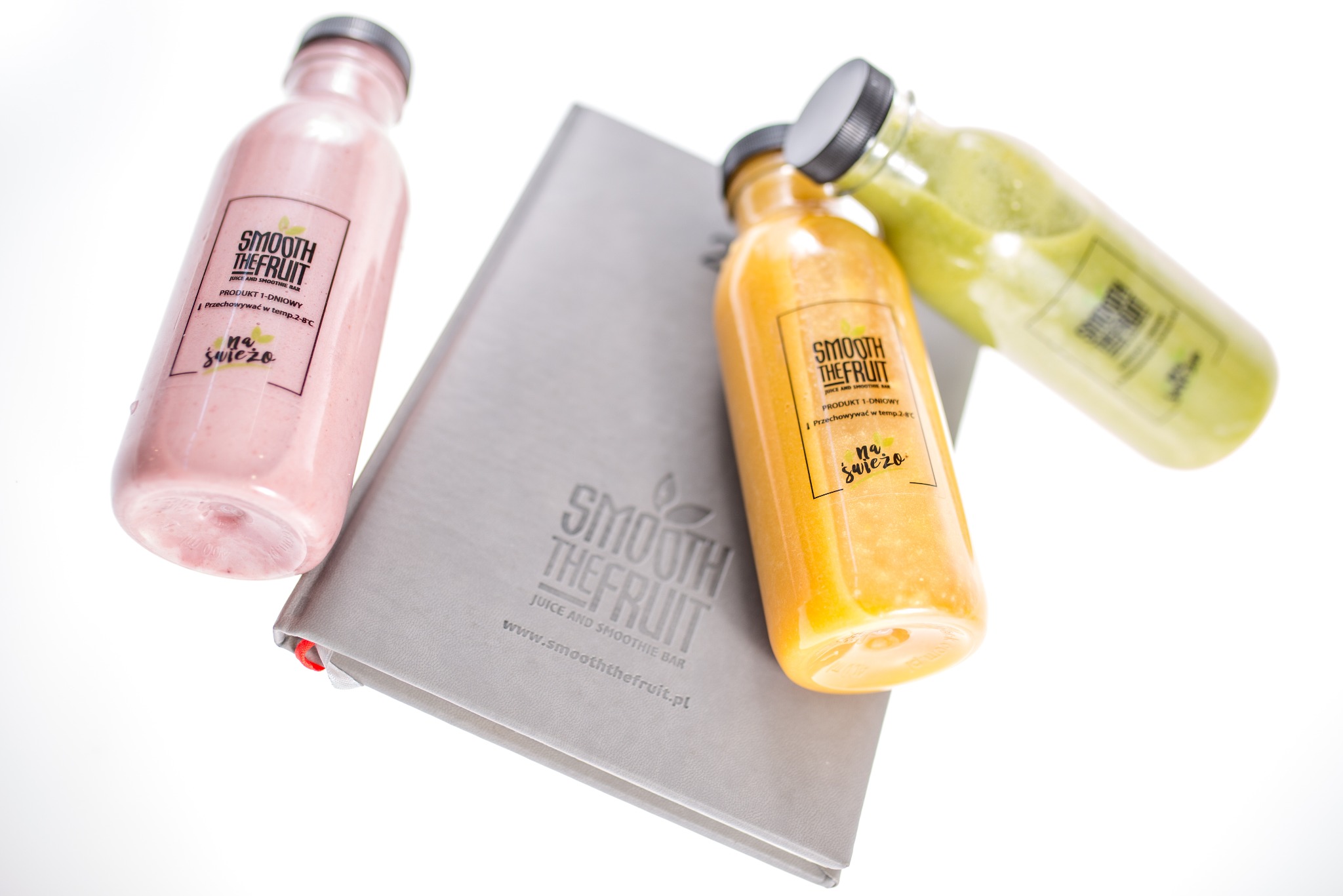 Trzy butelki świeżych soków i smoothie (różowy, pomarańczowy i zielony) z logo Smooth The Fruit, leżące na szarym menu tej samej marki, prezentowane na białym tle.