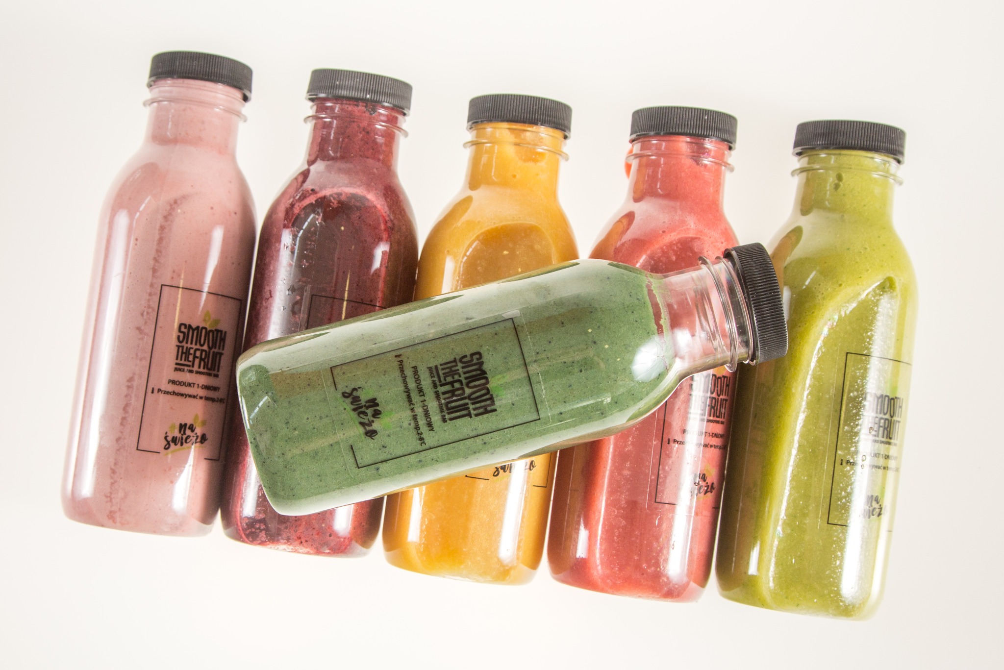Rząd plastikowych butelek z kolorowymi smoothie, w tym leżąca butelka z zielonym koktajlem, wszystkie z czarnymi zakrętkami i etykietami 'Smooth The Fruit'.