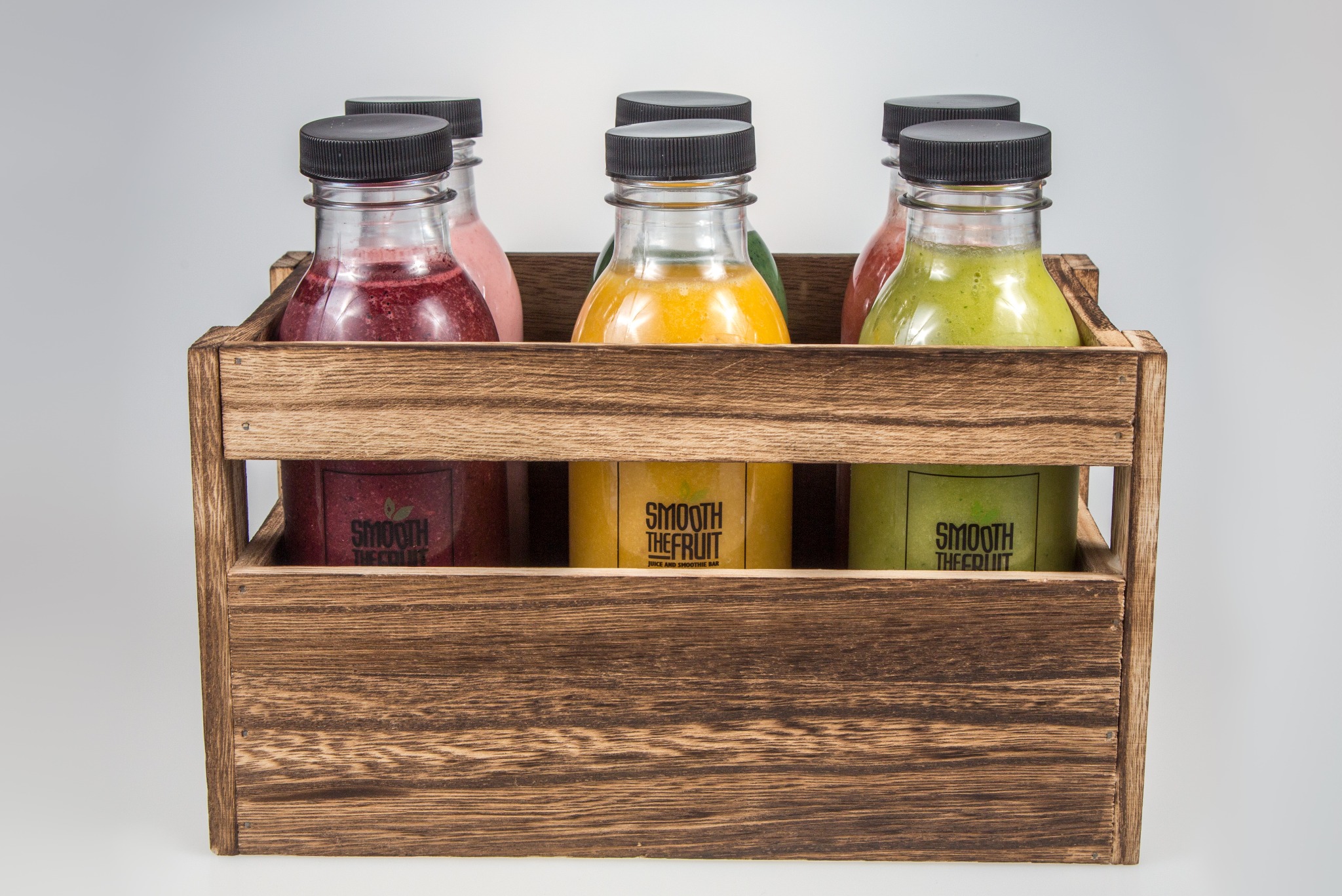 Sześć butelek ze świeżymi sokami w różnych kolorach, umieszczonych w drewnianej skrzynce z logo 'Smooth The Fruit', na białym tle.