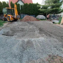 Żółta koparko-ładowarka obok pryzmy ziemi i rozłożonego żwiru na terenie budowy, w tle widać dom i zieleń, mężczyzna zagęszcza podłoże ubijarką.