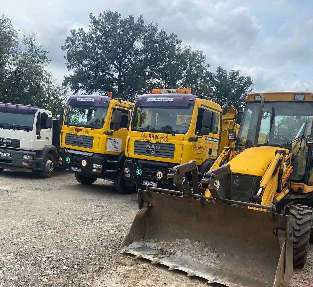 Rząd żółtych ciężarówek MAN z logo RRW i hasłem #nietakdrogadodroga oraz biała ciężarówka MAN i koparko-ładowarka na placu budowy, widok z przodu na łyżkę koparki, pochmurne niebo w tle.