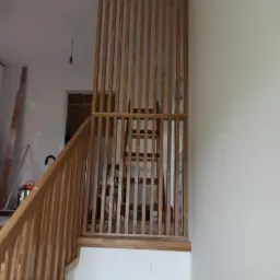 Drewniana balustrada z pionowych listew oddzielająca schody od antresoli, widoczna drabina w tle, surowy stan wykończenia wnętrza.