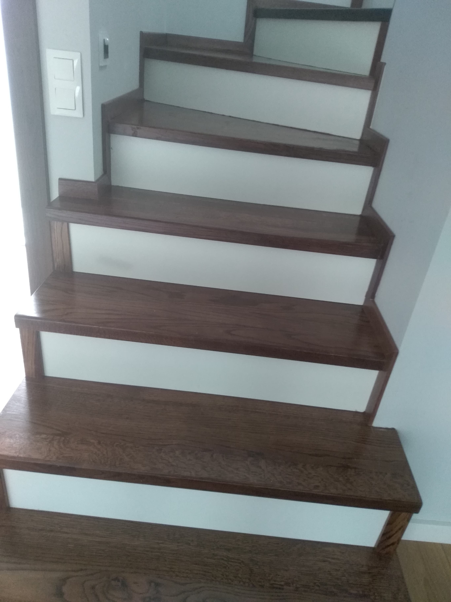 Drewniane schody z ciemnego drewna i białymi podstopnicami, widok z dołu, z widocznym włącznikiem światła na ścianie.