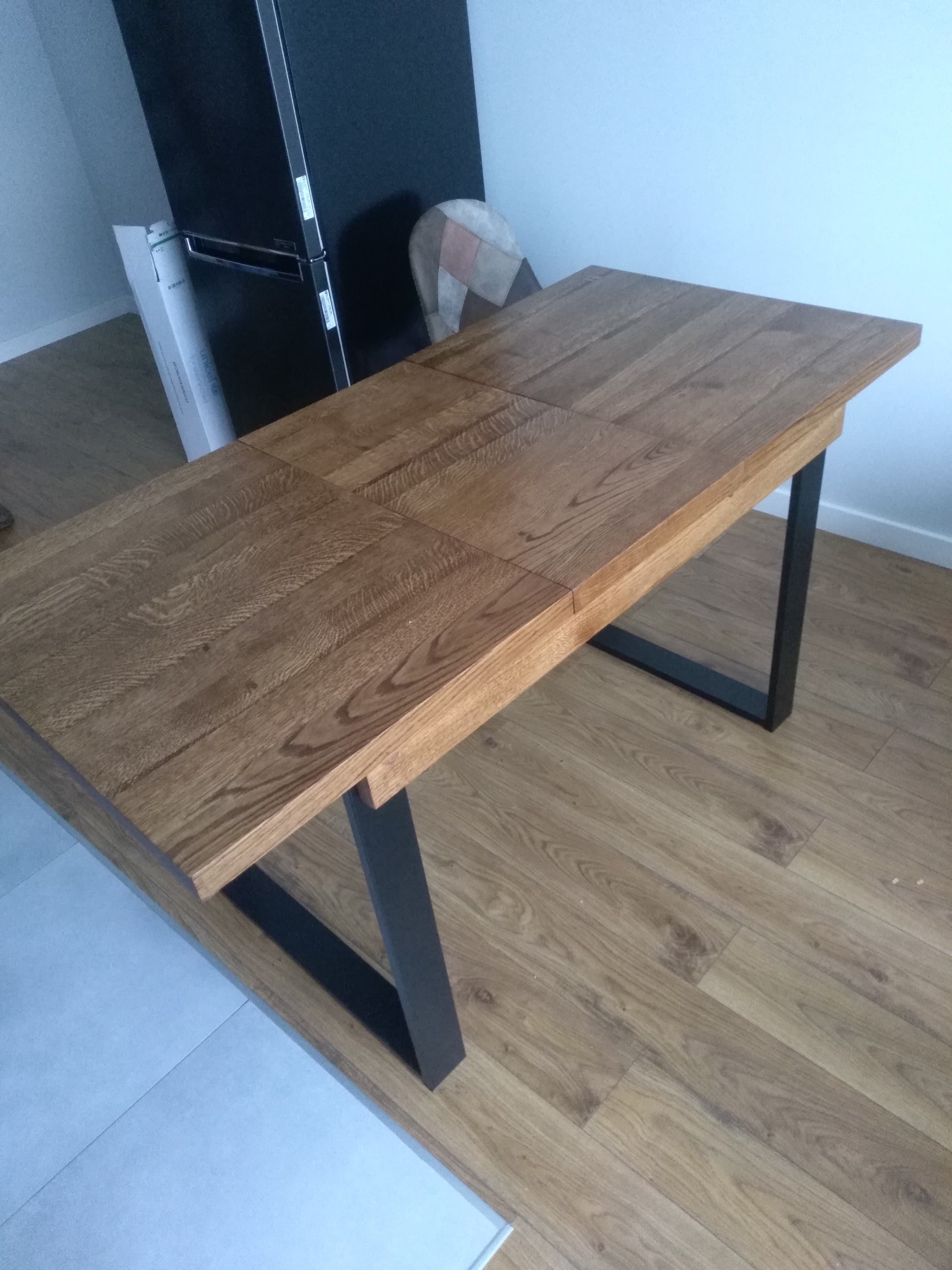 Rozkładany stół z blatem z drewna w odcieniach brązu, z czarnymi, metalowymi nogami w kształcie litery U, na podłodze z paneli imitujących drewno.