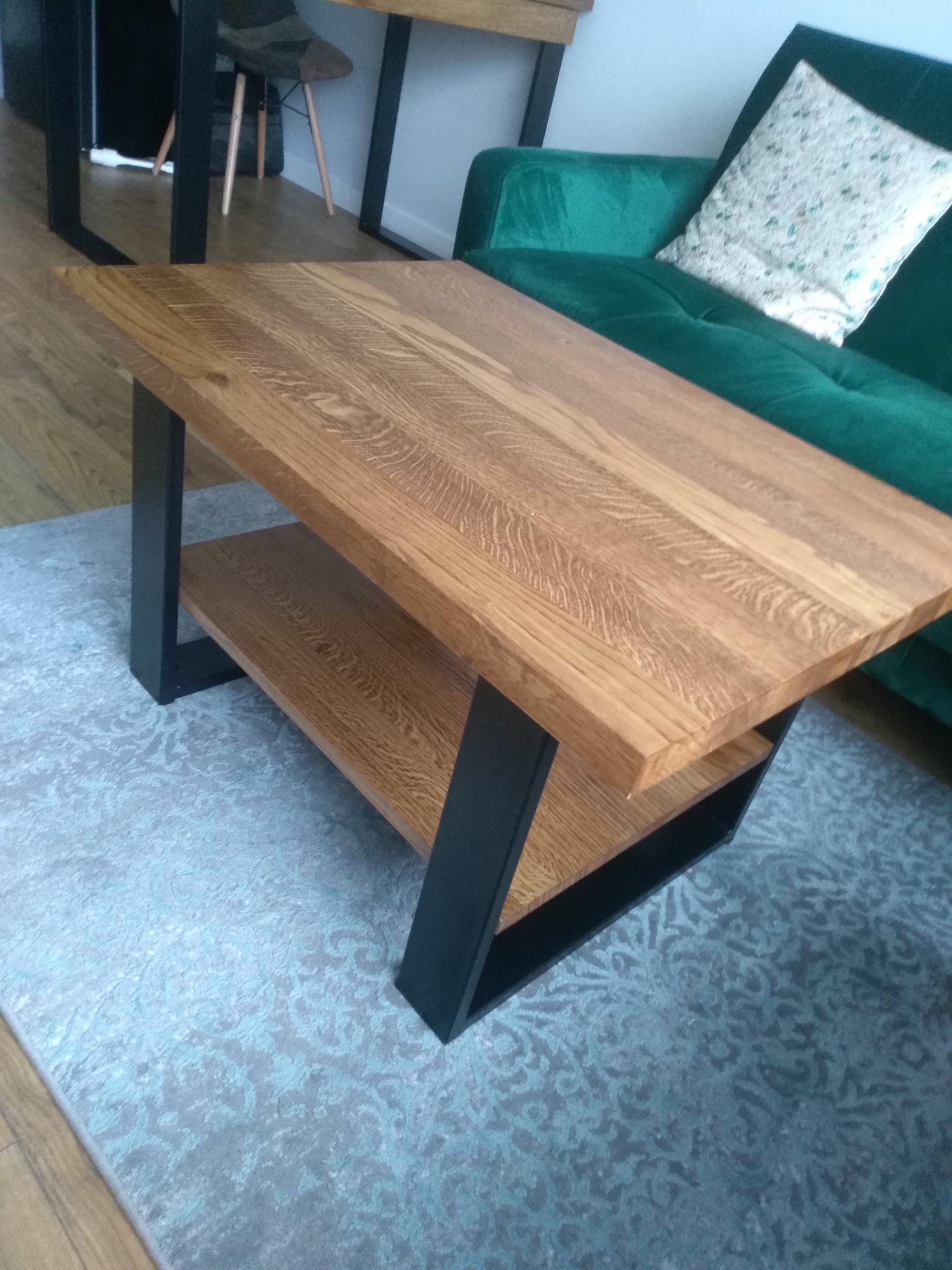 Stolik kawowy z jasnego drewna o wyraźnym usłojeniu, z dolną półką i czarnymi, metalowymi nogami, na tle zielonej sofy i wzorzystego dywanu.