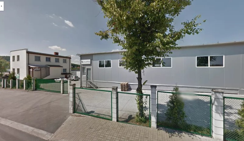 Budynek firmy z szarymi panelami, widoczny znak BASPOL, paleta drewniana obok wejścia, ogrodzenie z zieloną siatką.