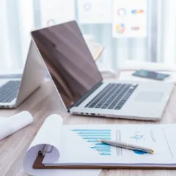 Dwa laptopy, dokumenty z wykresami i długopis na biurku w jasnym biurze.