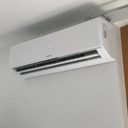 Biały klimatyzator Sinclair zamontowany na szarej ścianie w pomieszczeniu, temperatura wyświetlana na urządzeniu to 17 stopni, widoczny fragment karnisza nad urządzeniem.