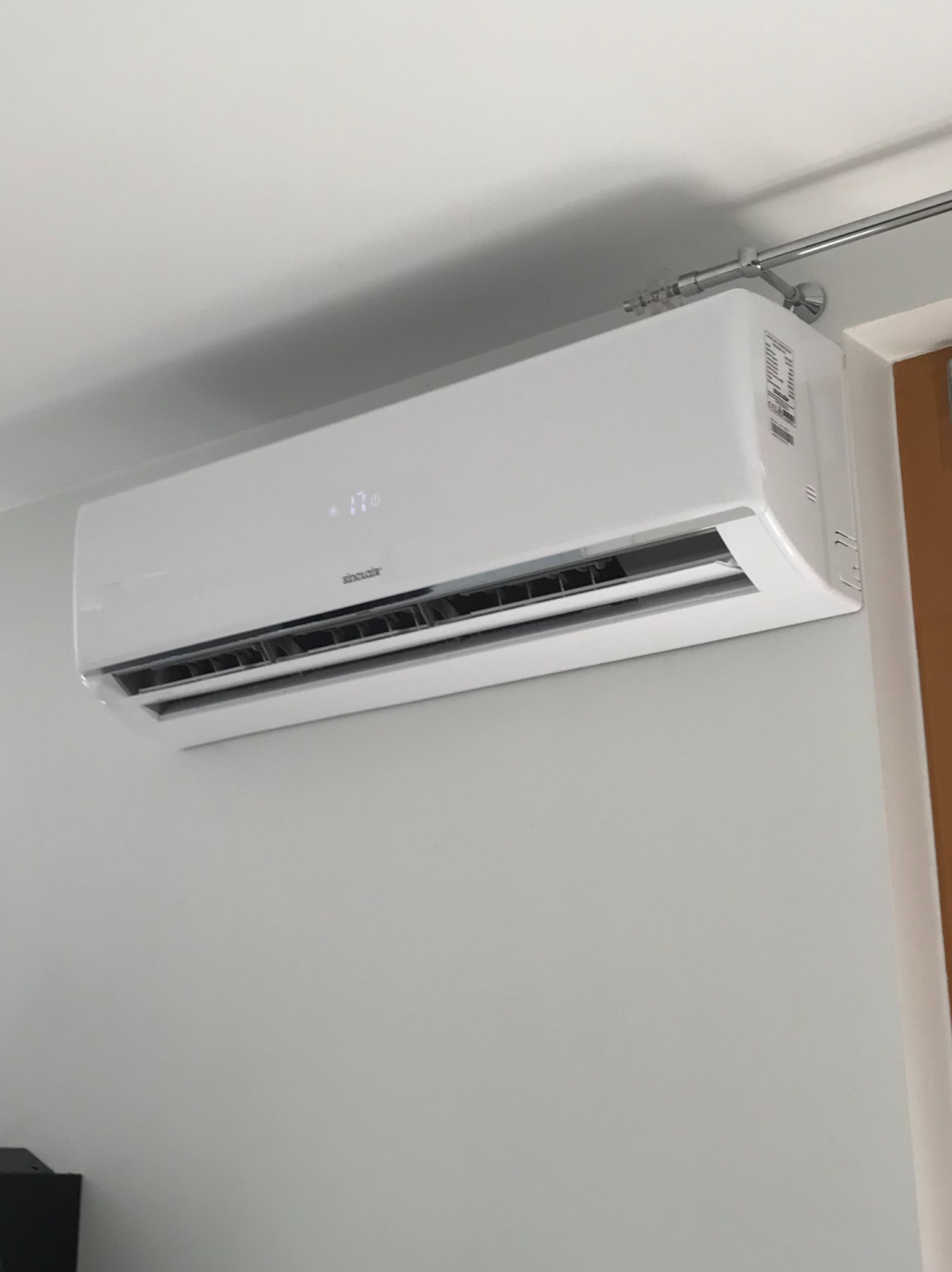Biały klimatyzator Sinclair zamontowany na szarej ścianie w pomieszczeniu, temperatura wyświetlana na urządzeniu to 17 stopni, widoczny fragment karnisza nad urządzeniem.