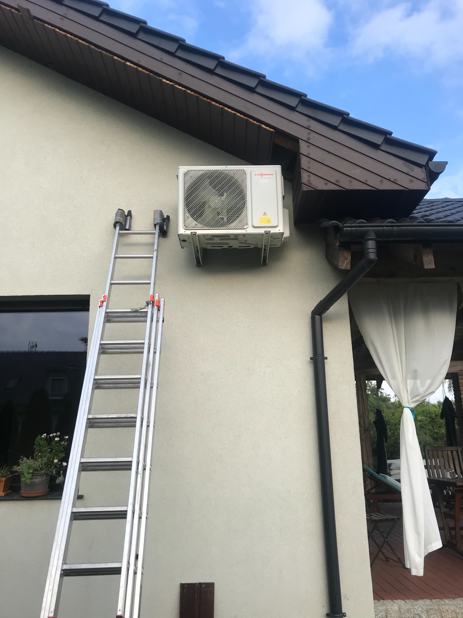 Zewnętrzna jednostka klimatyzacji Viessmann zamontowana na ścianie domu, drabina oparta o ścianę, widoczny fragment dachu i rynna.