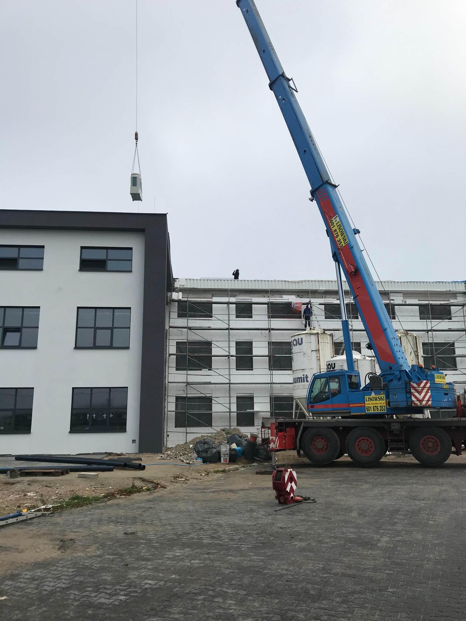 Montaż rekuperatora na dachu budynku komercyjnego za pomocą dźwigu, widoczne rusztowania i pracownicy na dachu.