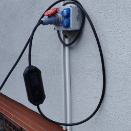 ELROM Firma Elektryczna Roman Oźmina - Zewnętrzna instalacja elektryczna z gniazdem siłowym i ładowarką EV, zamontowana na elewacji budynku. Widoczne kable i zabezpieczenia.