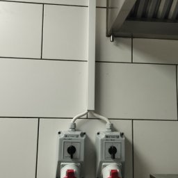 ELROM Firma Elektryczna Roman Oźmina - Dwa gniazda siłowe z czerwonymi wtykami, zamontowane na białej, kafelkowej ścianie. Nad nimi biała listwa maskująca przewody, a w tle fragment okapu kuchennego.