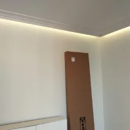 Pokój z zamontowanym oświetleniem LED w listwie przysufitowej, w tle kartonowe opakowanie IKEA oparta o ścianę i biała szafka.