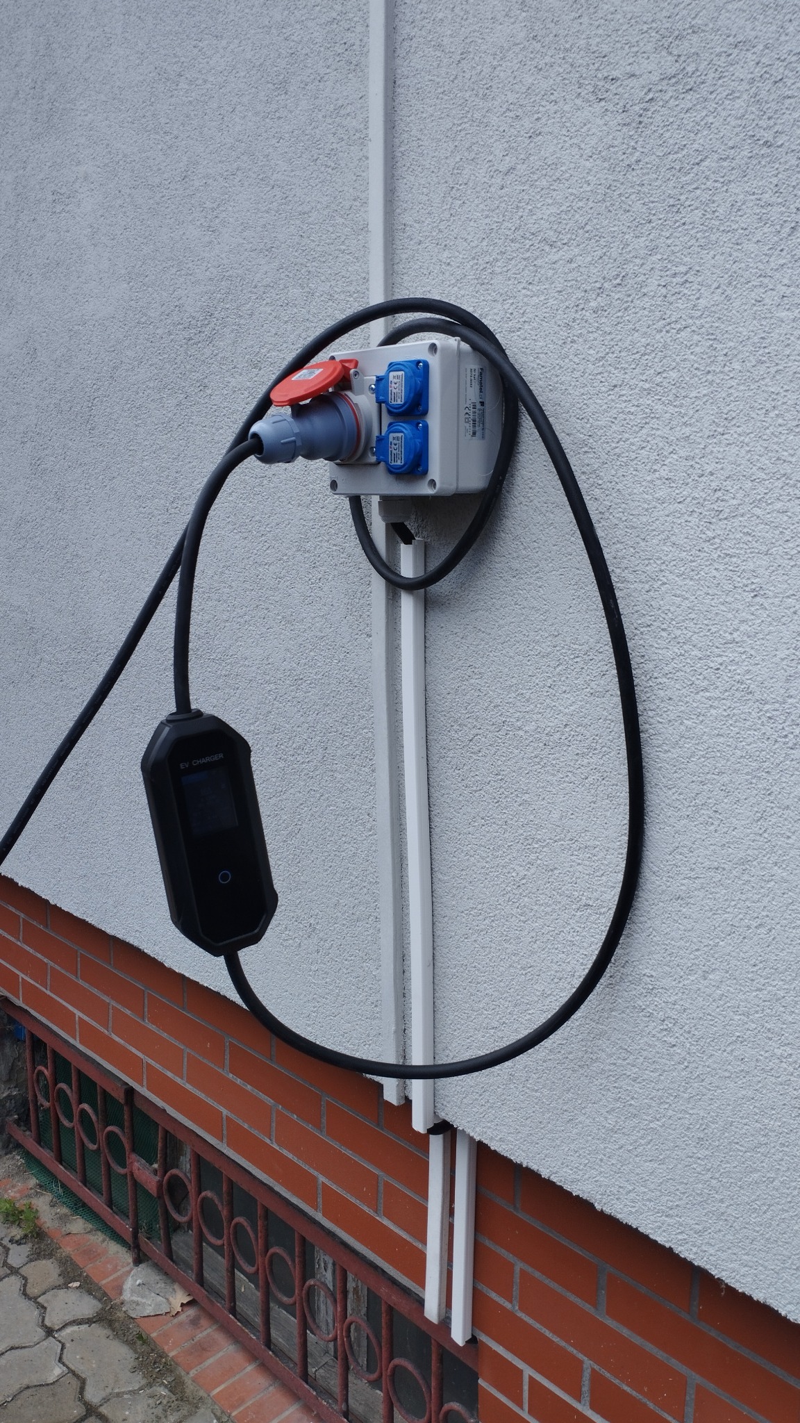 Zewnętrzna instalacja elektryczna z gniazdem siłowym i ładowarką EV, zamontowana na elewacji budynku. Widoczne kable i zabezpieczenia.