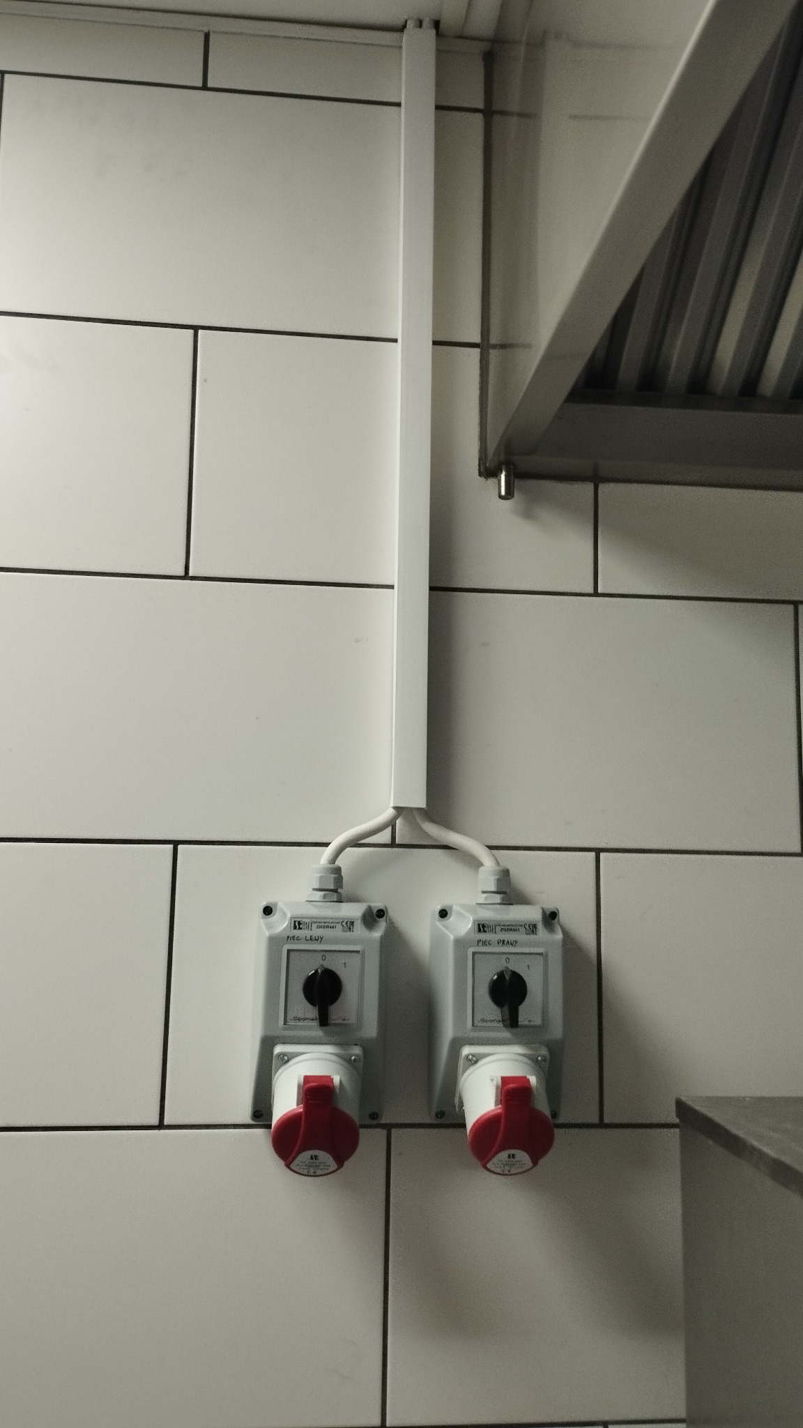Dwa gniazda siłowe z czerwonymi wtykami, zamontowane na białej, kafelkowej ścianie. Nad nimi biała listwa maskująca przewody, a w tle fragment okapu kuchennego.