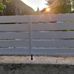 Nowoczesna, biała brama wjazdowa z poziomych profili, zamontowana między betonowymi słupkami, na tle zieleni i drewnianego budynku w promieniach zachodzącego słońca.