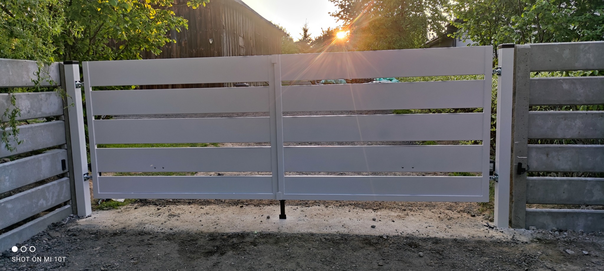 Nowoczesna, biała brama wjazdowa z poziomych profili, zamontowana między betonowymi słupkami, na tle zieleni i drewnianego budynku w promieniach zachodzącego słońca.