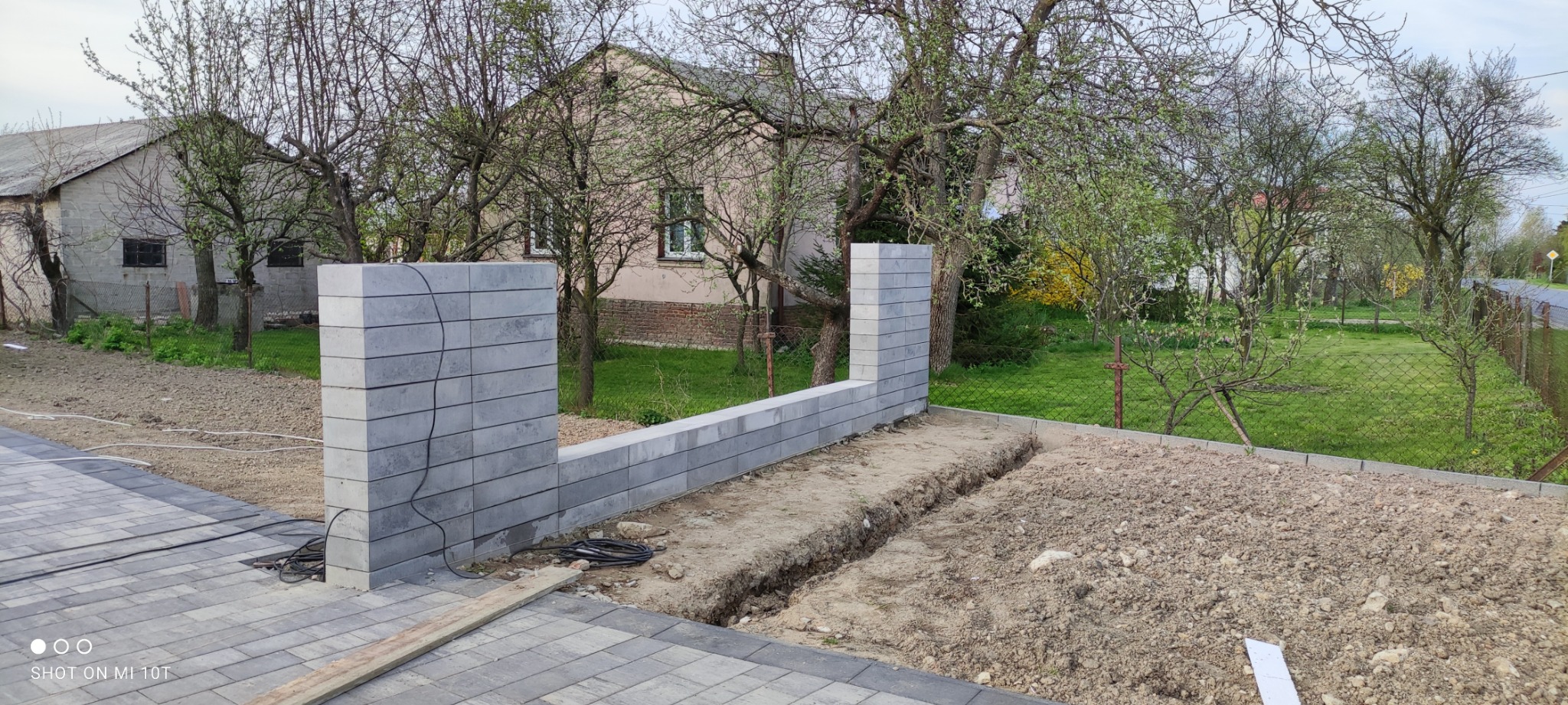 Wznoszenie murku z szarych betonowych bloczków, widoczny wykop pod fundament, w tle dom jednorodzinny i drzewa z wiosennymi pąkami.