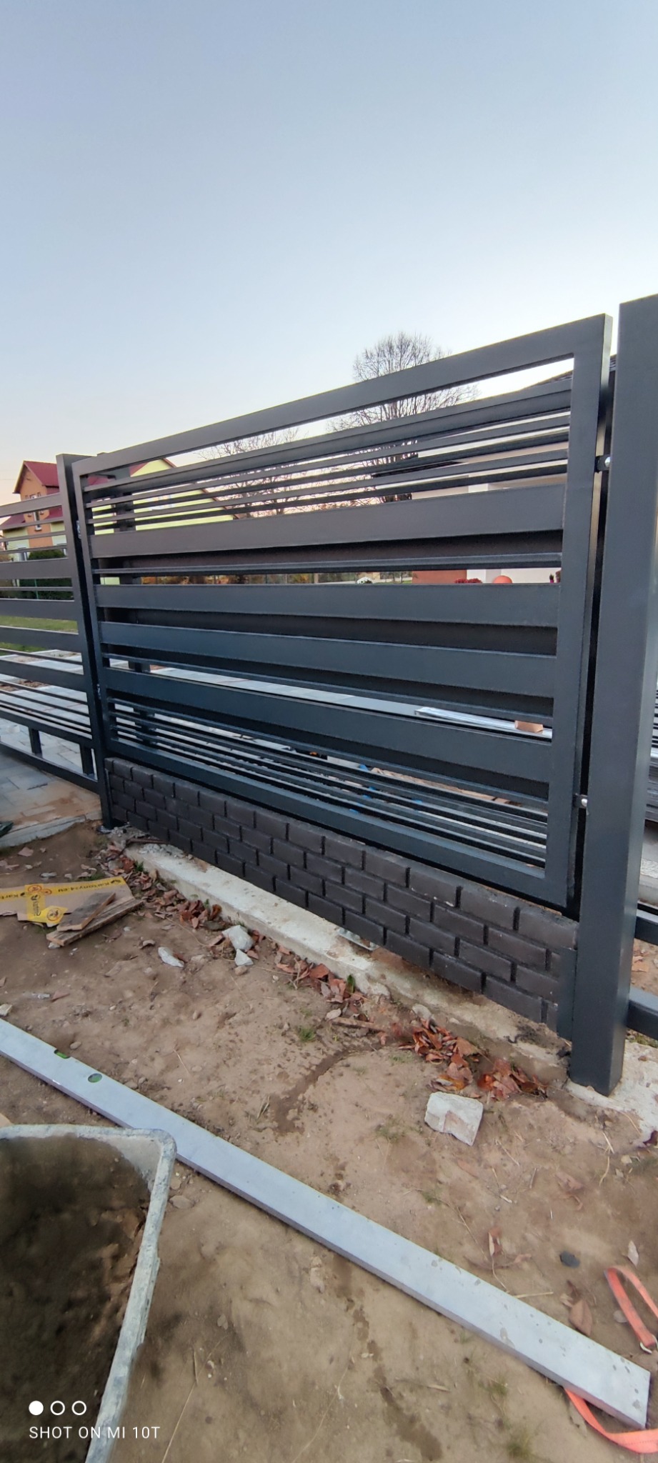 Nowoczesne ogrodzenie z poziomych, metalowych profili, osadzone na cokole z ciemnej cegły klinkierowej, w trakcie montażu, widoczne narzędzia i materiały budowlane.