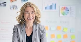 Uśmiechnięta kobieta w szarej marynarce, stojąca przed białą tablicą z wykresami i notatkami na samoprzylepnych kartkach, prezentująca koncepcję finansową.