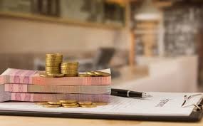 Stos banknotów przewiązanych gumką, kilka ułożonych w piramidkę złotych monet i czarny długopis leżące na dokumencie przypiętym do podkładki z klipsem, w tle rozmyte wnętrze pomieszczenia.