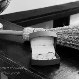 Czarno-białe ujęcie dwóch obrączek ślubnych w pudełku z cytatem, leżących obok miotły na lakierowanym drewnianym stole, z odbiciem w szklanej misie w tle.