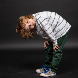 Portret chłopca z kręconymi włosami, pochylonego do przodu na czarnym tle, ubranego w koszulę w paski i zielone spodnie sztruksowe.