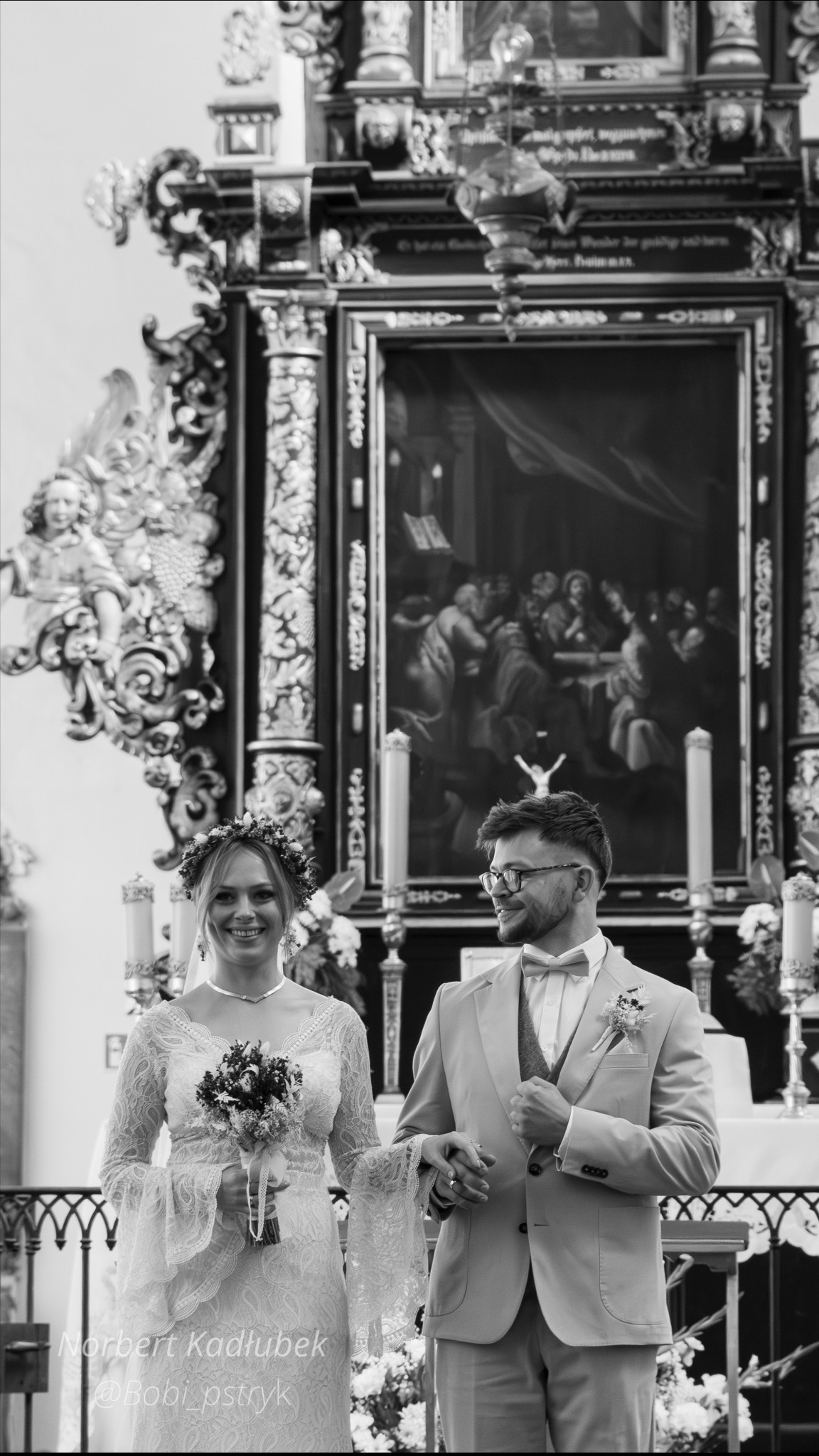 Czarno-białe zdjęcie pary młodej w kościele, panna młoda w koronkowej sukni i wianku, pan młody w jasnym garniturze i muszce, w tle ołtarz z obrazem.